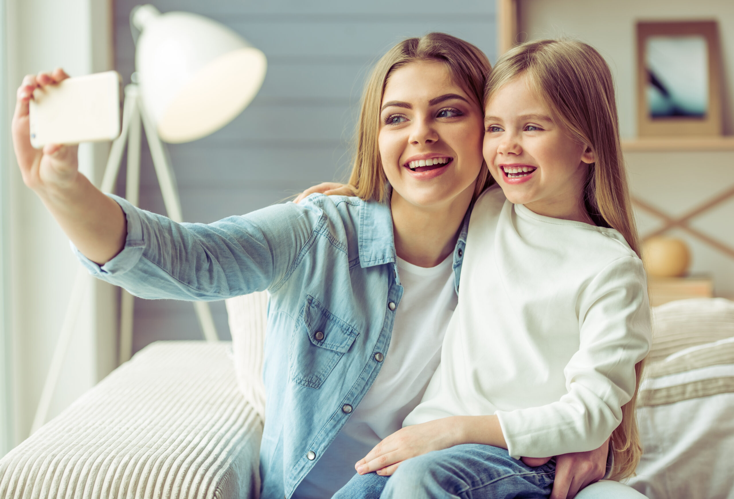 Beautiful young mom and her little daughter are making a selfie using a smart phone and smiling while sitting on sofa at home